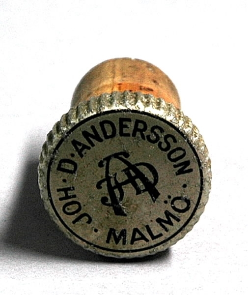 Flaska för hårvatten. a) Flaska med tre beige tätt sammanklistrade etiketter. Vänster etikett: Blomsterslingor, brunt tryck, text: "Petral hårvatten verksammaste hårtvättmedel till aflägsnande af mjäll och skadliga mikrober utan risk för förkylning". Mittetiketten: Kvinna i halvfigur hållande en ros, brunt tryck, text: "Petral hårvatten Joh. D. Andersson Malmö". Höger etikett: Namnchiffer, brunt tryck, text: "Petral hårvatten förhindrar alla hårsjukdomar, stärka hårrötterna och befodra hårväxten. Joh. D. Andersson, Malmö". Ovanför etiketterna en liten lapp, text: "Torr". På motstående sida längst ned lapp, text: "Farligt att förtära". Runt flaskhalsen etikett, text: "Petral hårvatten Joh. D. Andersson Malmö". b) Kork. Etikett med namnchiffer, text: "John. D. Andersson Malmö".