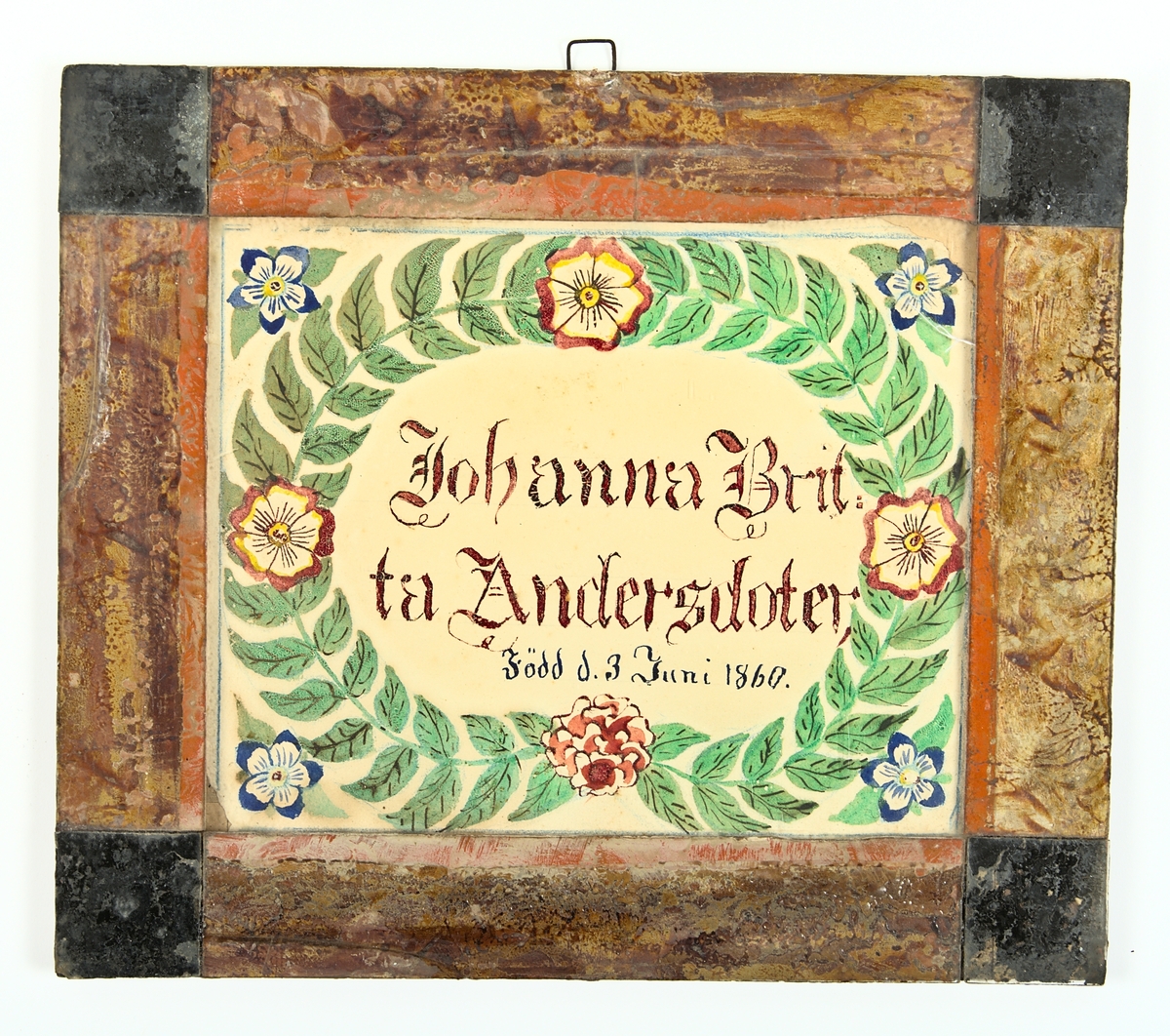 Minnestavla, glasad och inramad med målade glasbitar, imiterande en mahognyram med ebenholtshörn. Text och målning på papper. Text: Johanna Brit:ta Andersdotter Född d. 3 Juni 1860. inom en krans av gröna blad med 4 röda blommor. I vardera hörnet, utanför kransen, en blå blomma.