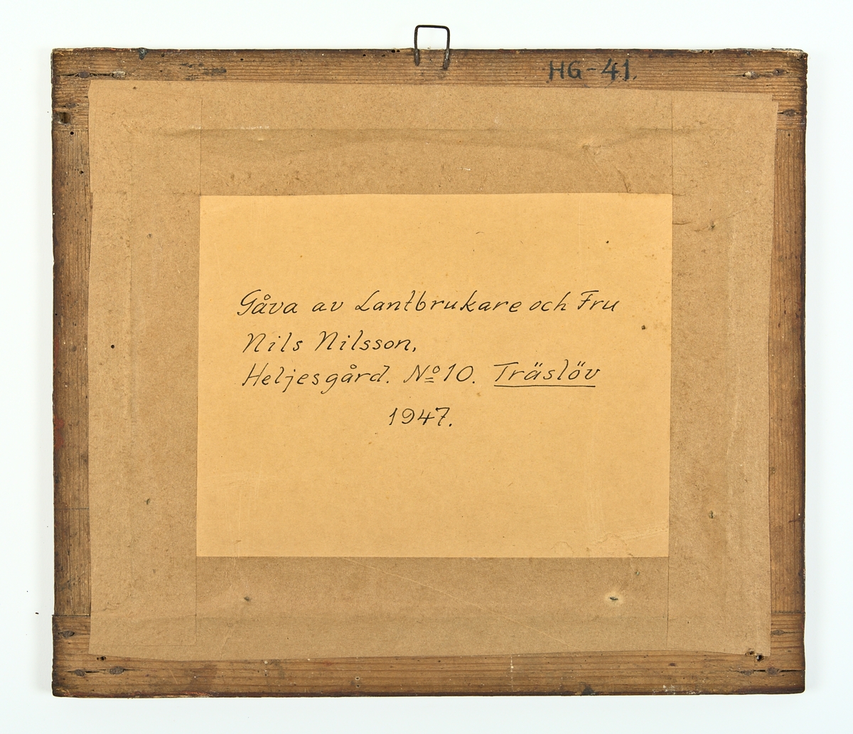 Minnestavla, glasad och inramad med målade glasbitar, imiterande en mahognyram med ebenholtshörn. Text och målning på papper. Text: Johanna Brit:ta Andersdotter Född d. 3 Juni 1860. inom en krans av gröna blad med 4 röda blommor. I vardera hörnet, utanför kransen, en blå blomma.