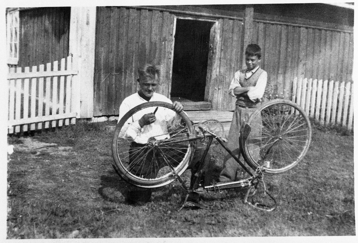 Hestsveen nordre på Vennevold, Østre Toten, ca. 1935. Bilde tatt på sydsida av husmannsstua med den låge inngngsdøra. Kristoffer Bern reparerer sykkelen sin med Erling Kristiansen som tilskuer.