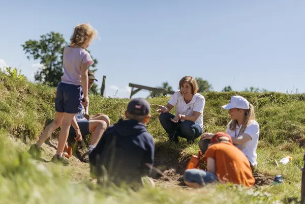 Barn leker ute i gresset, noen sitter og noen står. Det er sol