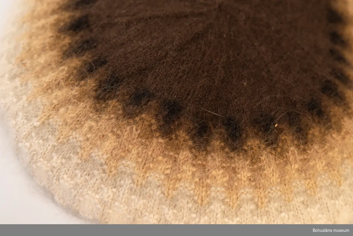 Stickad barett med slätstickade enfärgade partier med en resårstickad kant. Baretten är mönsterstickat med räta och aviga maskor i mörkbrunt, svart, ljusbrunt, brunbeige, grått och vitt mot en brun botten.