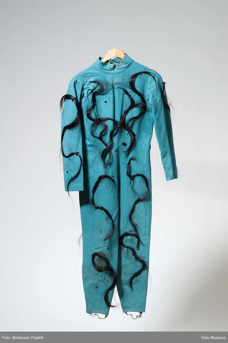 Turkisfarget hel drakt i crepe- påsydde, sorte hårdusker. Lukkes med glidelås i ryggen.