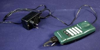 :1 Mobiltelefon bestående av rektangulärt hölje av mörkgrön och svart hårdplast i två delar som sitter ihop, inuti vilken telefonapparatens elektronik finns. På ena sidan ett antal knappar av grått rspektive svart gummi på vilka siffror och tecken står tryckt med vit text: siffrorna 0-9, stjärna, fyrkant, pil, rund ring med prick i, samt en telefonlur med pil som visar att en lur läggs av. Knapparna används när man ringer med telefonen genom att slå abbonentens telefonnummer eller när man vill använda någon annan telefonfunktion. Ovanför knapparna hål för högtalare där man sätter örat för att höra den man pratar med. Under knapparna hål för mikrofon där man pratar under samtalet. Vit tryckt text ovan- och under knapparna: "Personal Phone" samt "Motorola" och en logotype med ett M inom en cirkel. På andra sidan sitter telefonens batteri av rektangulärt format metallstycke med svart plasthölje, vilket kan tas loss och sättas dit. På vit klisterlapp står med svart tryckt text: "Motorola 6V Nickel - cadmium battery" mm, samt symbolen för återvinningsbar: tre pilar. Orange prislapp med priset 398:- fastklistrad på batteriet.
:2 Batteriladdare bestående av dosa av plast i vilken en stickkontakt sitter. Denna sticks in i ett elektriskt vägguttag vid laddning. Genom en svart ca 1500 mm lång sladd som i ena änden sitter i dosan och i andra änden har en stickkontakt av plast som sätts i ett uttag i telefonen, förs ström till telefonbatteriet så att det laddas.