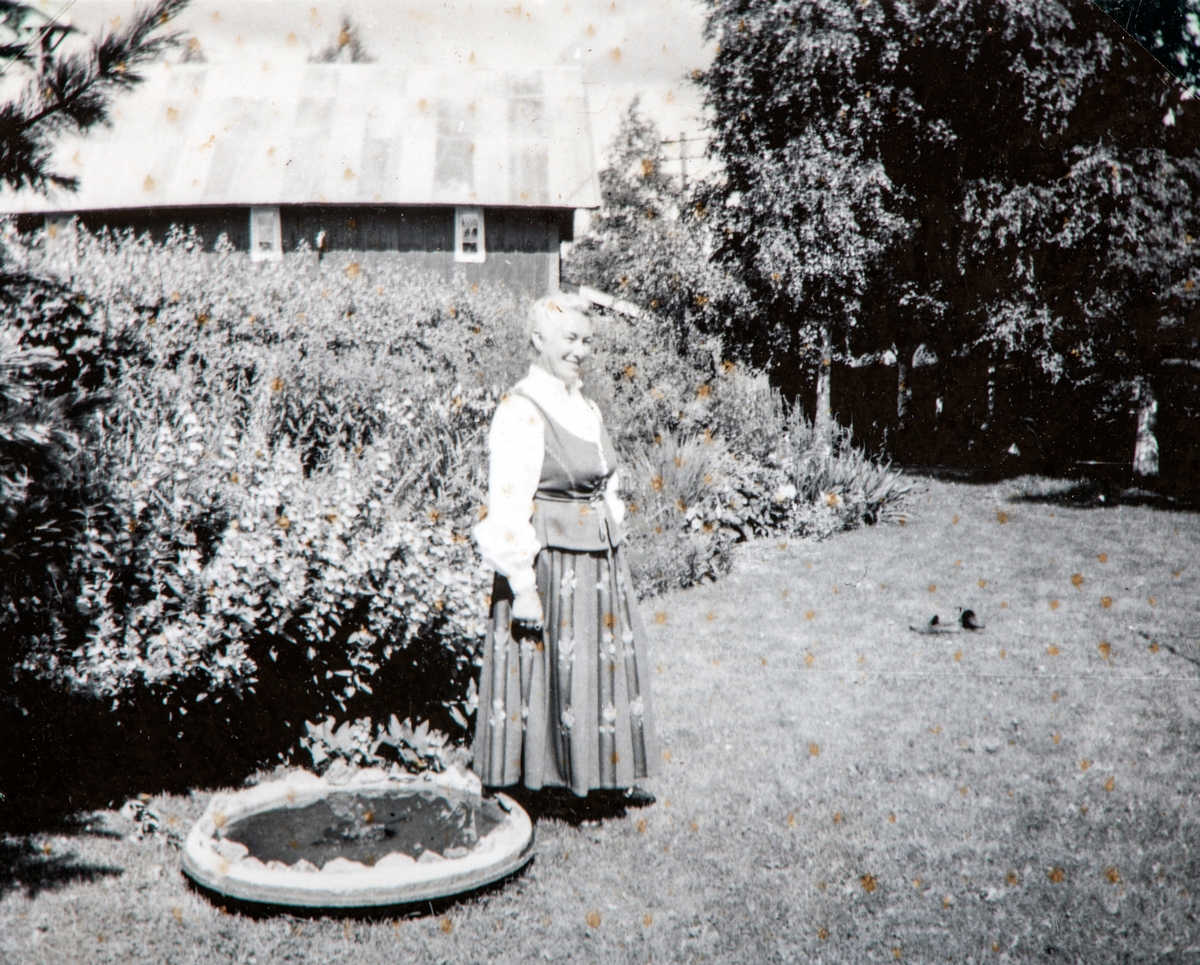 Oddrun Storbæk i hagen på Grylling, Løten, ca midt på 1950-tallet. Staudebed, fuglebad. Hedmarksbunad.