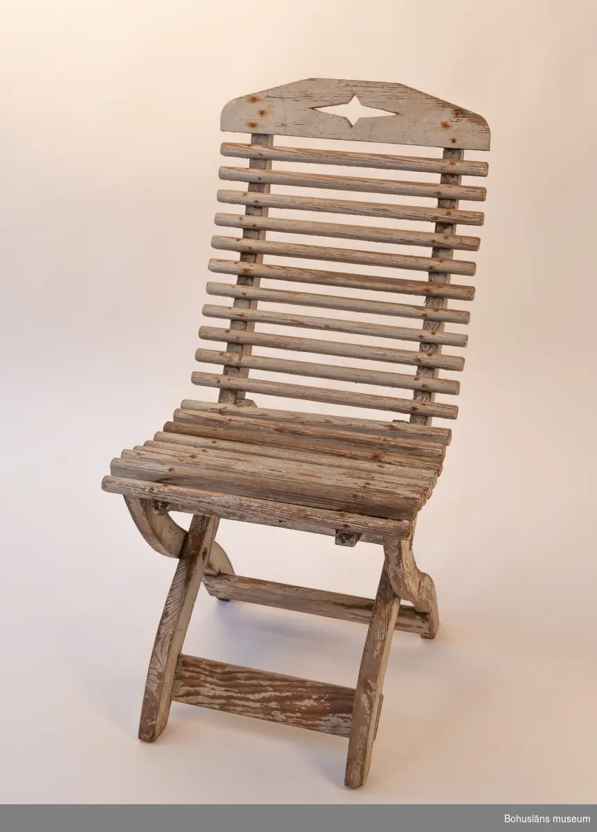 Vitmålad trästol, hopfällbar. Sits av rundade kraftiga spjälor. Dekorativt utformade benavslut och ryggbricka med utsågad stjärnform.
Samhör med UM032292 Bord.