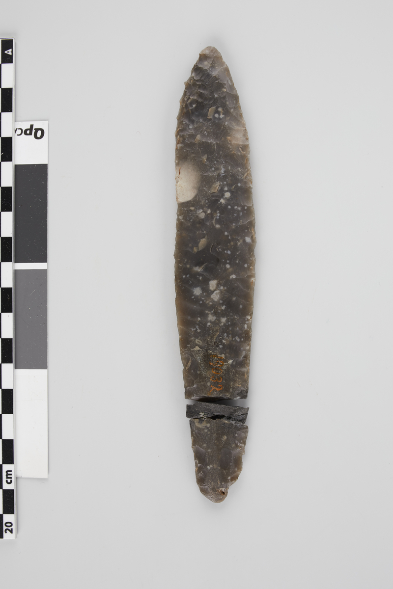 Spydspids af Flint, som NO. 68; har været et smukt, vel arbeidet Exemplar, men er brudt nær nedre Ende, og et lidet Stykke mangler i Bruddet. Oprindelig Længde c. 18 cm., største Bredde 3,3 cm.