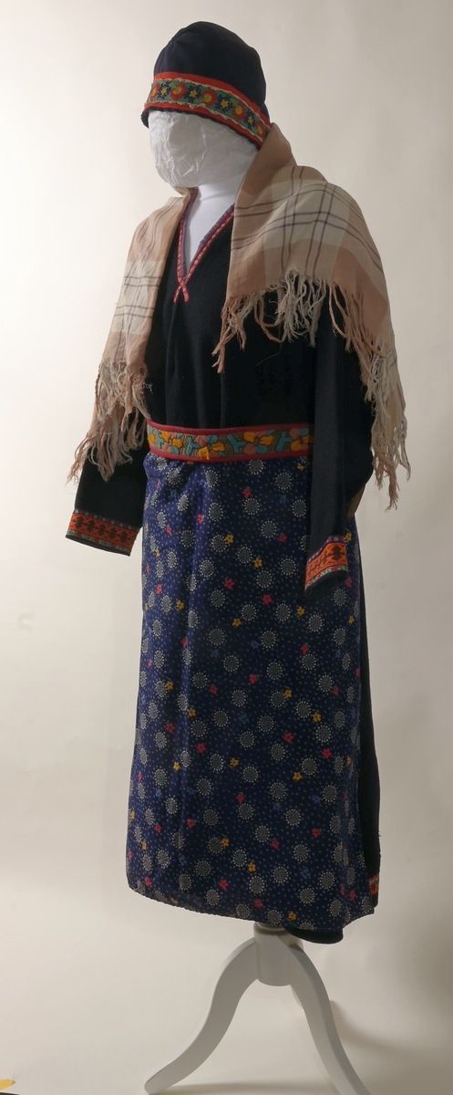 Kofte med lue, belte, forkle og sjal. Lua har dekorbånd nederst langs rimmen. Selve kofta har vevde dekorasjonsbånd på jakkeermene, kragen, oppe på og langs skuldrene, og nederst på kofta. Beltet til kofta låses ved lærreim som tres inn i lås med låsepinne - alt av stål. Selve låsen skjules av beltet da lærreim og lås er på baksiden av beltet. Beltets ende er avsluttet av tynt flettet bånd. Båndet festes i tinnknapp som er på utsiden av beltet. Dette er ei dekorativ løsning for å skjule den funksjonelle stållåsen. Forklet er blått, og dekorert med hvite snøkrystaller og blomster. Blomstene er gule, røde og blå blomser.  Kofta er i svart som er hovedfargen på kofta såvel som lua, mens dekorbånd og lue har andre farger.