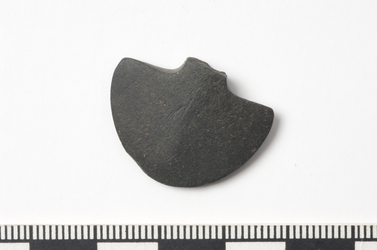 Fragment av et spadeformet hengesmykke av skifer, brukket ved overgangen til halsen. Stl.: 20 mm, stb.: 26 mm, stt.: 6 mm. 