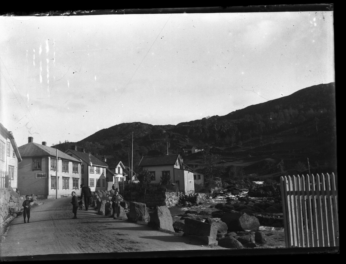 Barn i en gate med trehus. En elv renner forbi.