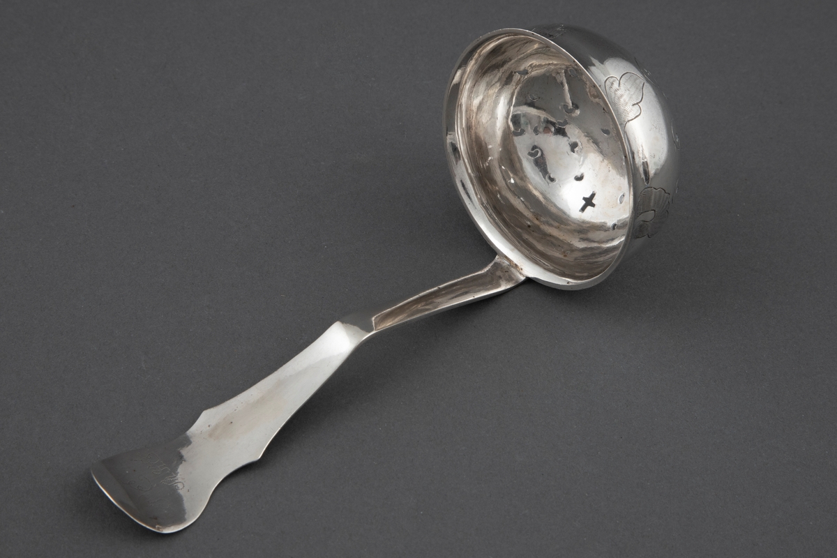 Sukkerstrøskje i sølv. Rundt, bolleformet perforert blad med svakt utoverbøyet rand. På skjebladets underside er det gravert stilisert blomsterdekor. Skaftroten er loddrett på bladet og får en knekk utover midt på skaftet, for så ved enden å bli bøyet noe innover igjen. Skaftets bredde tiltar mot enden som er avrundet. På bladkanten og skaftendens underside er det et gravert og punslet rokokko-ornament.