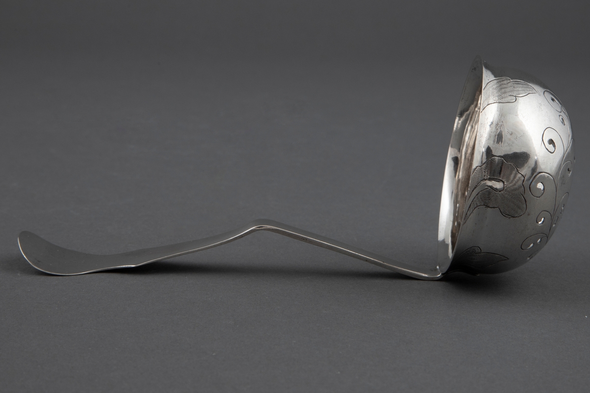 Sukkerstrøskje i sølv. Rundt, bolleformet perforert blad med svakt utoverbøyet rand. På skjebladets underside er det gravert stilisert blomsterdekor. Skaftroten er loddrett på bladet og får en knekk utover midt på skaftet, for så ved enden å bli bøyet noe innover igjen. Skaftets bredde tiltar mot enden som er avrundet. På bladkanten og skaftendens underside er det et gravert og punslet rokokko-ornament.