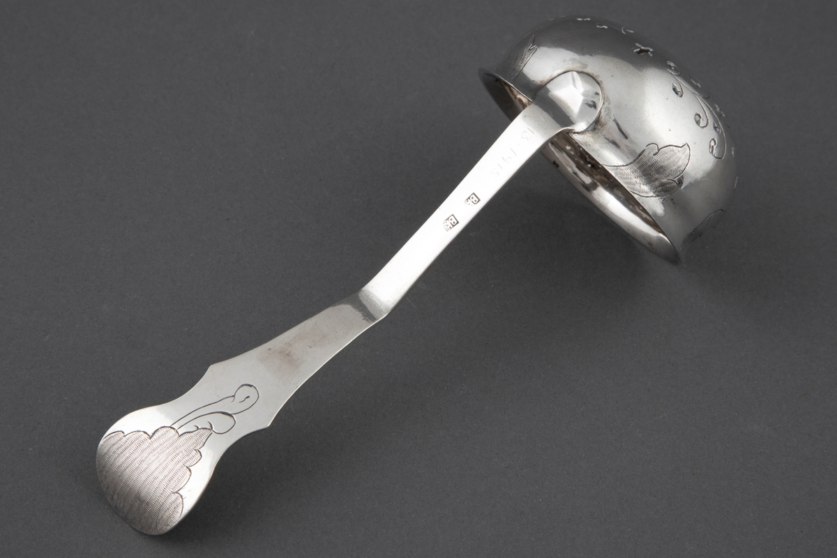 Sukkerstrøskje i sølv. Rundt, bolleformet perforert blad med svakt utoverbøyet rand. På skjebladets underside er det gravert stilisert blomsterdekor. Skaftroten er loddrett på bladet og får en knekk utover midt på skaftet, for så ved enden å bli bøyet noe innover igjen. Skaftets bredde tiltar mot enden som er avrundet. På bladkanten og skaftendens underside er det et gravert og punslet rokokko-ornament.