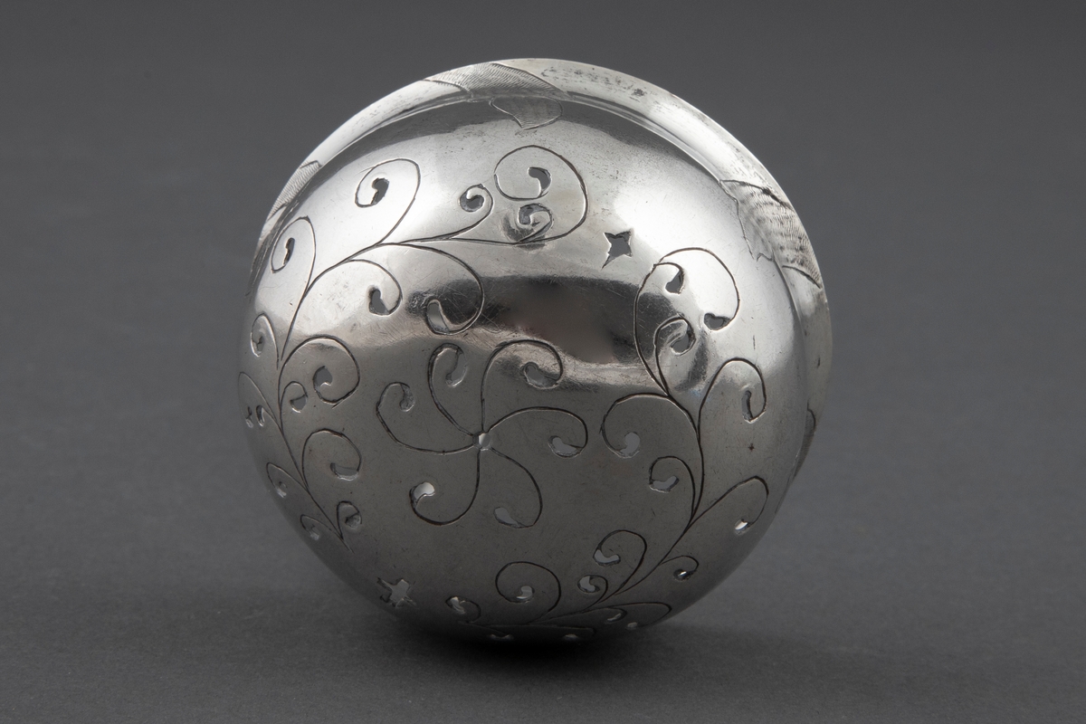 Sukkerstrøskje i sølv. Rundt, bolleformet perforert blad med svakt utoverbøyet rand. På skjebladets underside er det gravert stilisert blomsterdekor. Skaftroten er loddrett på bladet og får en knekk utover midt på skaftet, for så ved enden å bli bøyet noe innover igjen. Skaftets bredde tiltar mot enden som er avrundet. På bladkanten og skaftendens underside er det et gravert og punslet rokokko-ornament.