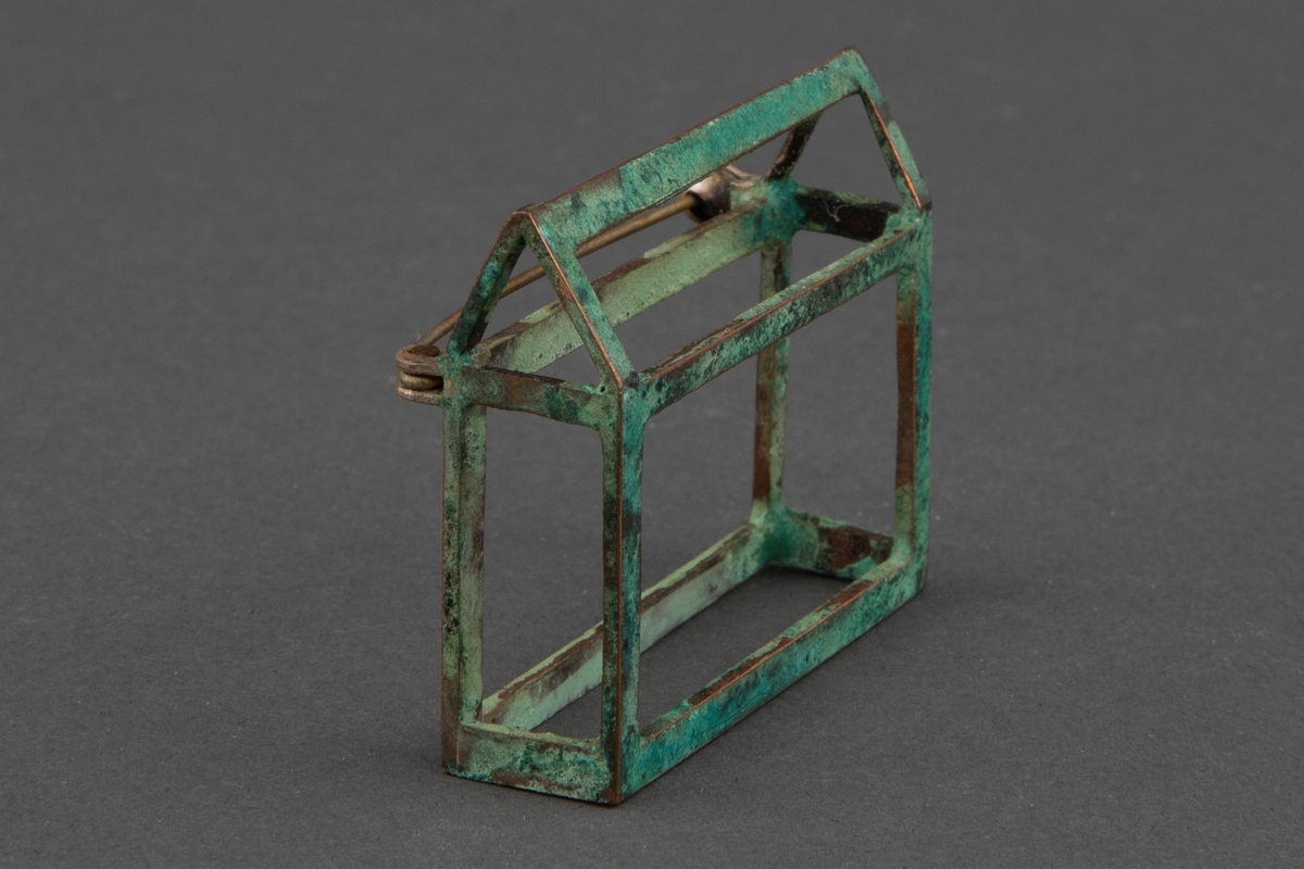 Brosje i patinert kobber. Brosjen har en arketypisk horisontal husform uten vegger.