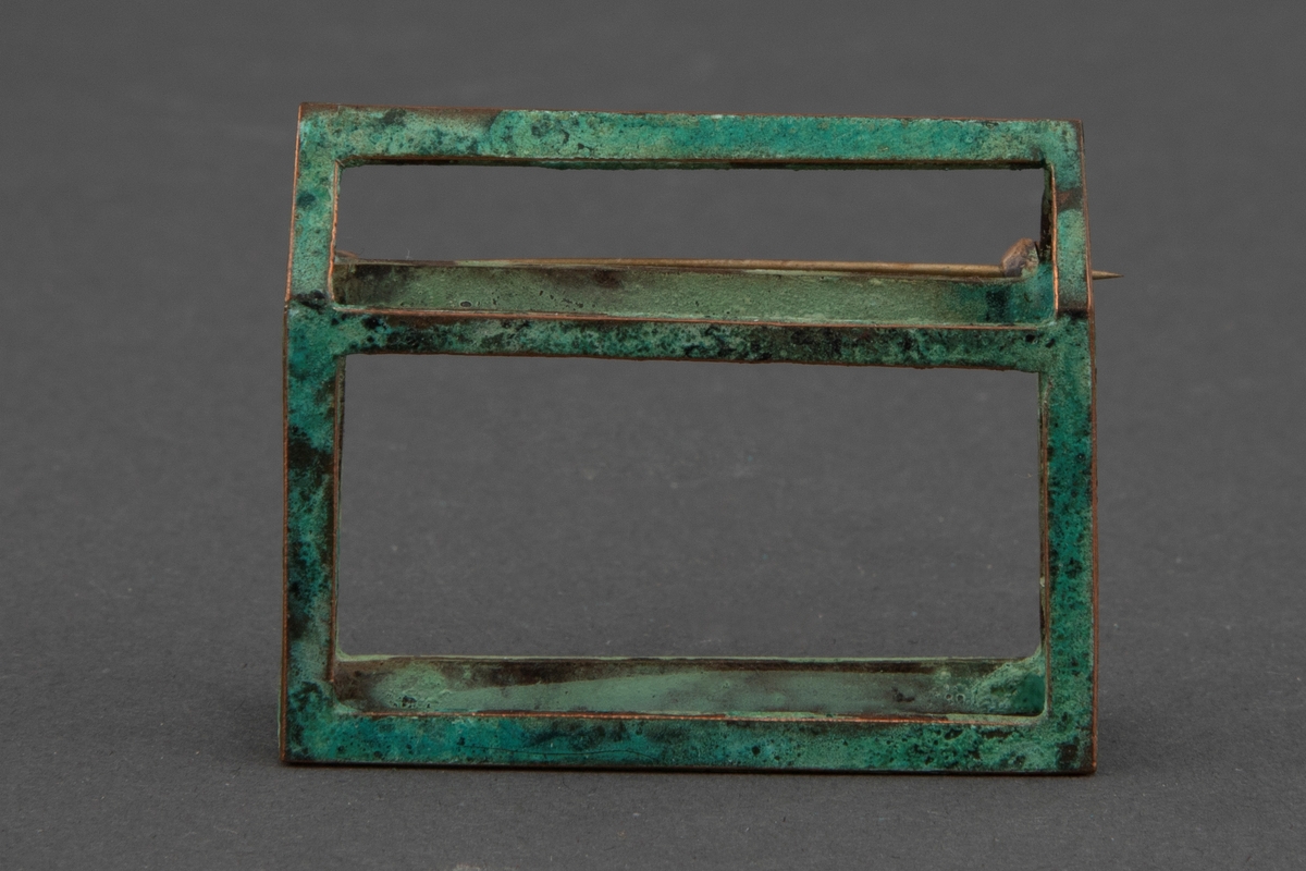 Brosje i patinert kobber. Brosjen har en arketypisk horisontal husform uten vegger.