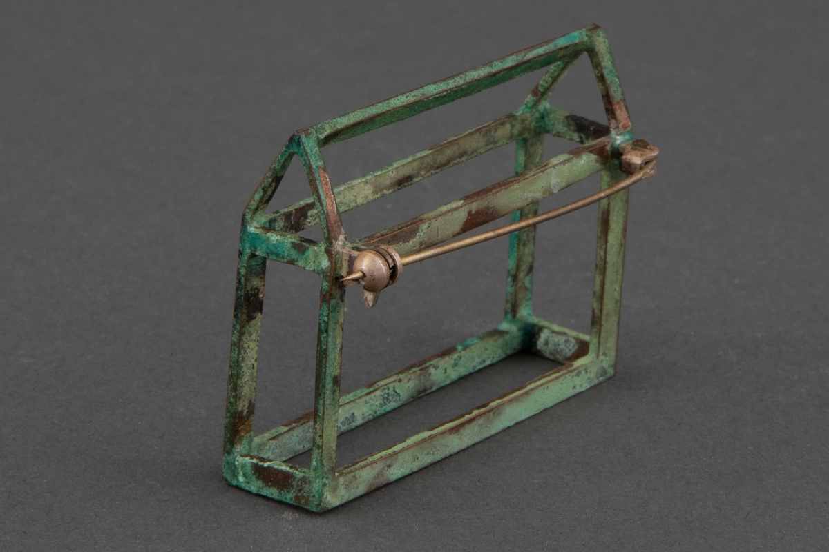 Brosje i patinert kobber. Brosjen har en arketypisk horisontal husform uten vegger.