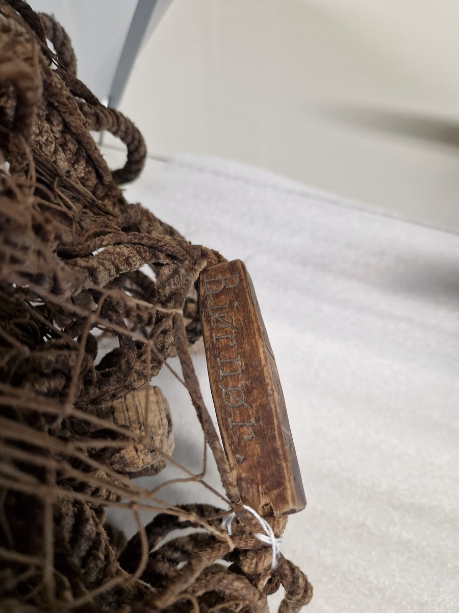 Lin og hampsnøre bunnet sammen til masker (garnlinet), påfestet korkebiter og gjennomhulete trebiter med innskåret bokstaver og tall