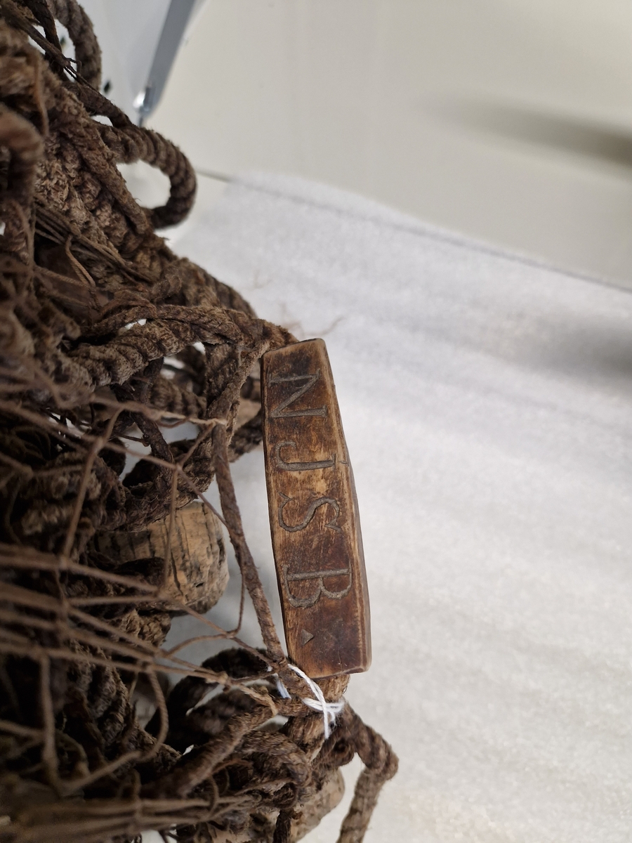 Lin og hampsnøre bunnet sammen til masker (garnlinet), påfestet korkebiter og gjennomhulete trebiter med innskåret bokstaver og tall