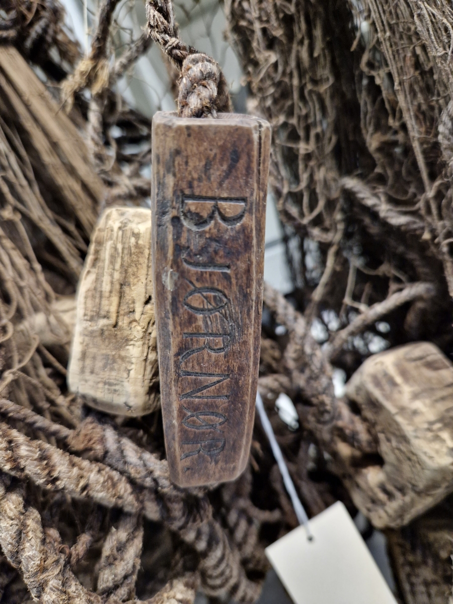 Lin og hampsnøre bunnet sammen til masker (garnlinet), påfestet korkebiter og gjennomhulete trebiter med innskåret bokstaver og tall
