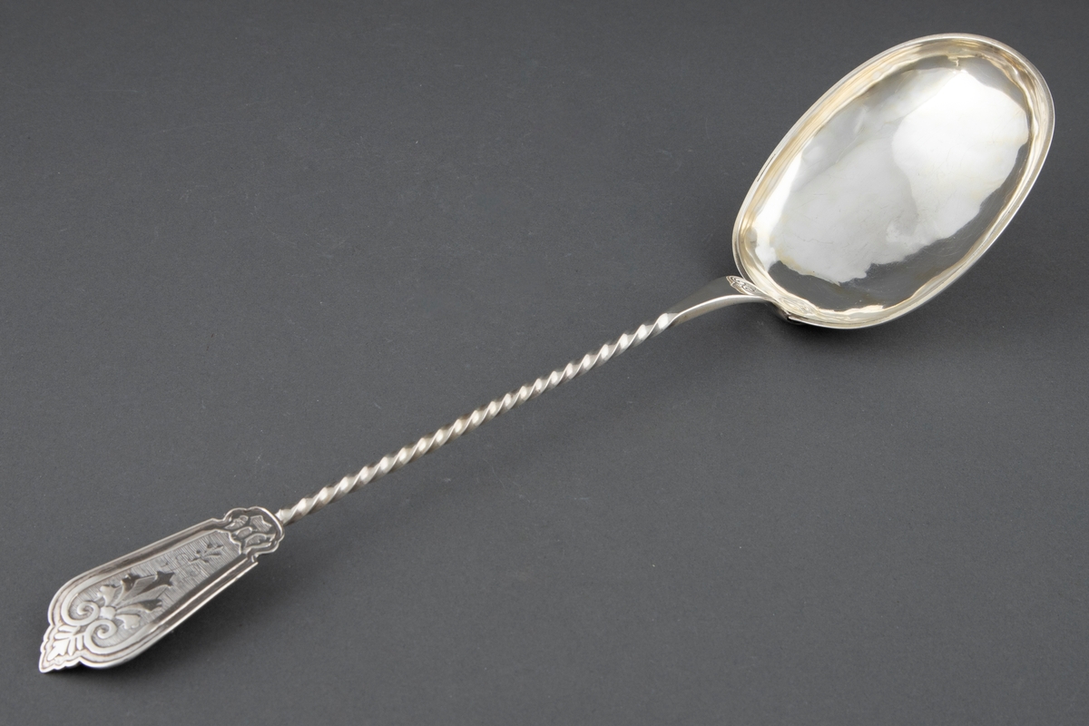 Suppeøse i sølv. Stort ovalt skjeblad med påloddet kant og innvendig forgylling. Tvunnet skaft som er avsluttet med en påloddet plate med pressede, vifteformede ornamenter på riflet bunn. Enkel gravert ornamentikk på overgangen til skjebladet.