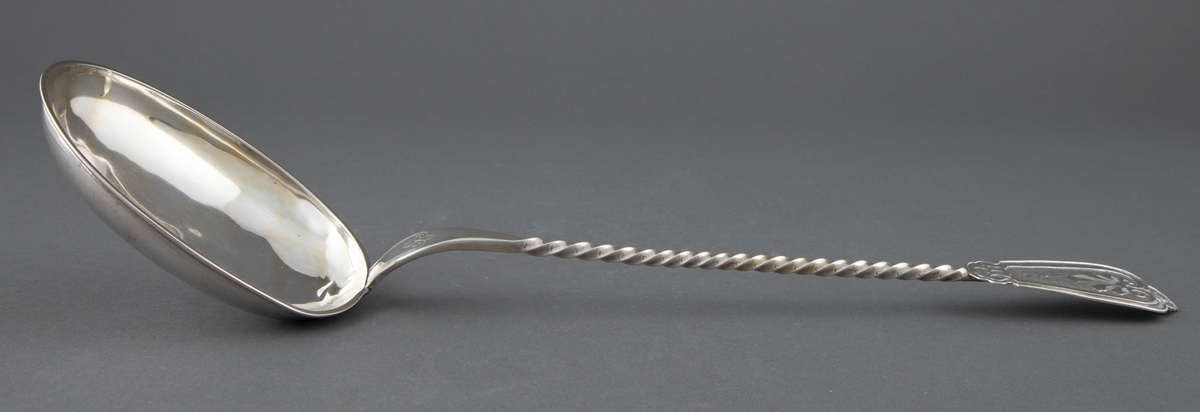 Suppeøse i sølv. Stort ovalt skjeblad med påloddet kant og innvendig forgylling. Tvunnet skaft som er avsluttet med en påloddet plate med pressede, vifteformede ornamenter på riflet bunn. Enkel gravert ornamentikk på overgangen til skjebladet.