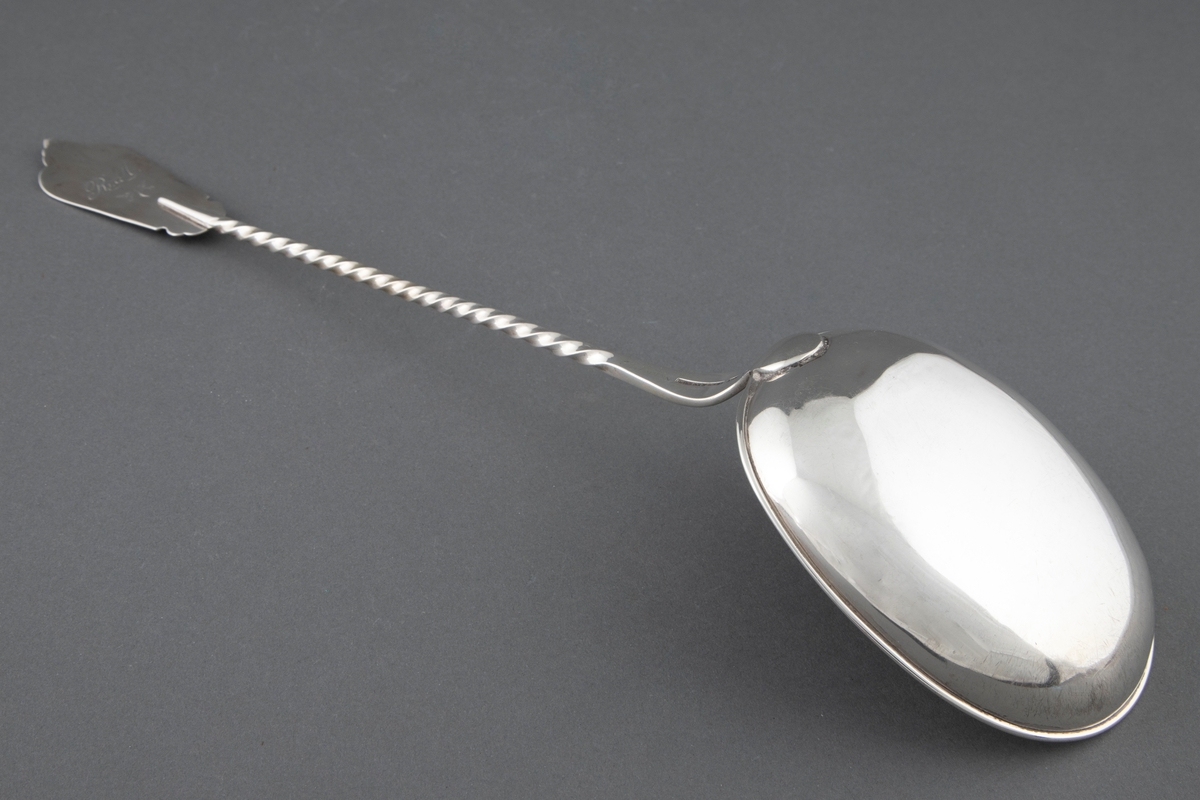 Suppeøse i sølv. Stort ovalt skjeblad med påloddet kant og innvendig forgylling. Tvunnet skaft som er avsluttet med en påloddet plate med pressede, vifteformede ornamenter på riflet bunn. Enkel gravert ornamentikk på overgangen til skjebladet.