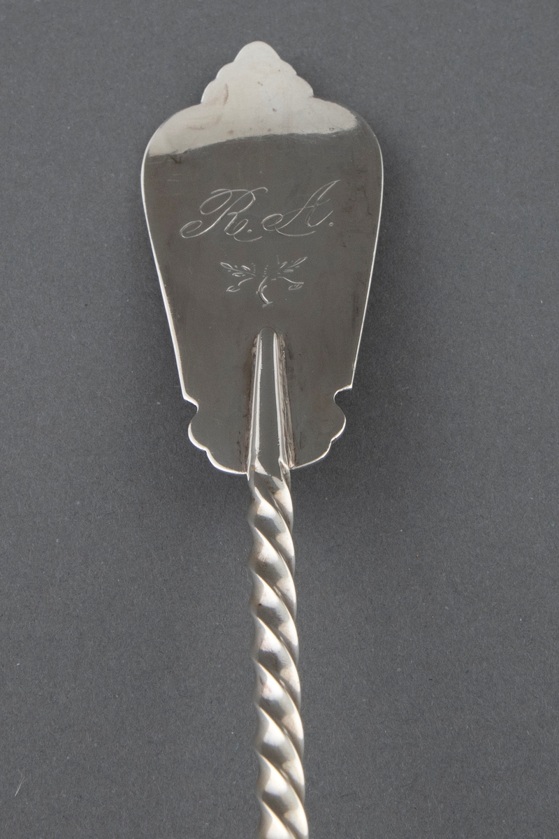 Suppeøse i sølv. Stort ovalt skjeblad med påloddet kant og innvendig forgylling. Tvunnet skaft som er avsluttet med en påloddet plate med pressede, vifteformede ornamenter på riflet bunn. Enkel gravert ornamentikk på overgangen til skjebladet.