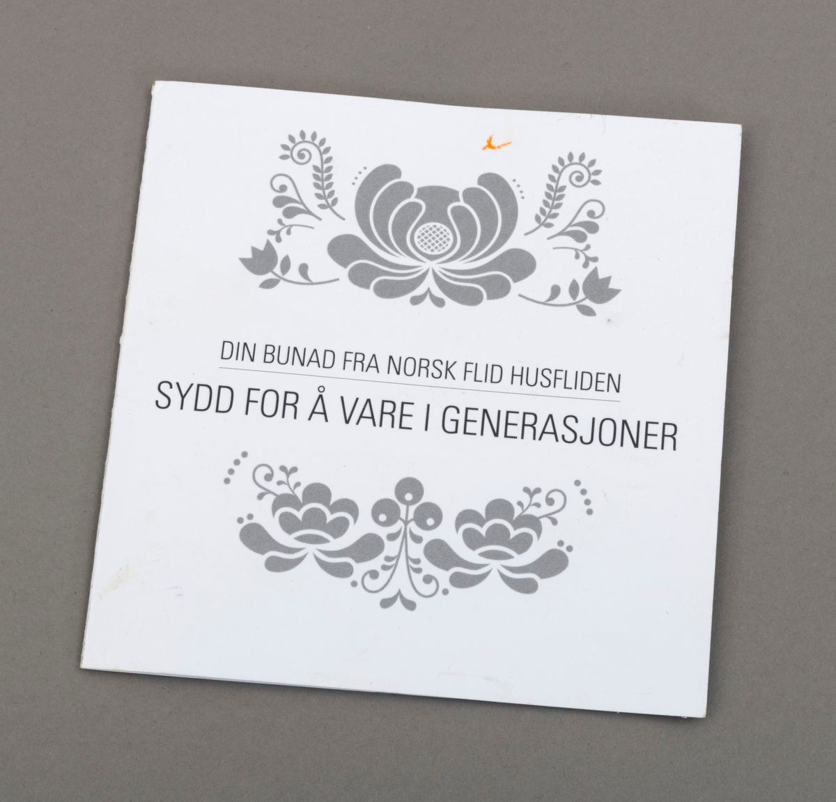 Gavekort fra Norsk Flid Husfliden "sydd for å vare i generasjoner". Hvit bakgrunne med sølvfarget akantusliknende dekor og påskrift. Kortet er sammenbrettet.