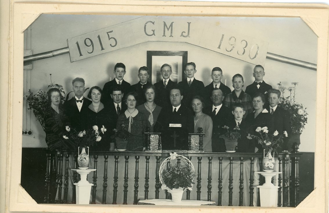 Gruppbild med vuxna och ungdomar uppställda bakom på ett podium under en skylt med texten: "1915 G M J 1930".