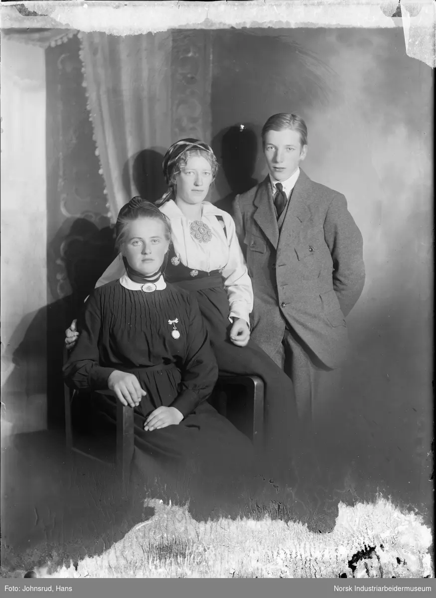 Gruppeportrett. To kvinner og en mann fotografert i studio. Den ene kvinnen sitter i en stol og er kledd i mørk kjole med smykkebånd rundt halsen og kyse med knytebånd under haka. Den andre kvinnen sitter på stolens ermlene og er kledd i folkedrakt med sølje på brystet og skaut på hodet. Mannen er kledd i dress med slips.