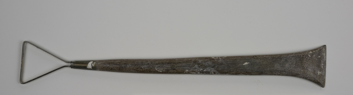 Modellerslynge brukt av Nils Aas. Slyngen består av et flatt metallbånd festet i enden av et trehåndtak. Motsatt ende av håndtaket er formet som en spatel. Det er noe rester etter gips.