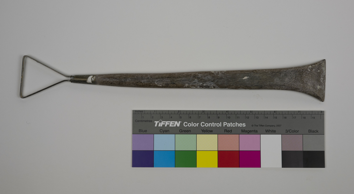 Modellerslynge brukt av Nils Aas. Slyngen består av et flatt metallbånd festet i enden av et trehåndtak. Motsatt ende av håndtaket er formet som en spatel. Det er noe rester etter gips.