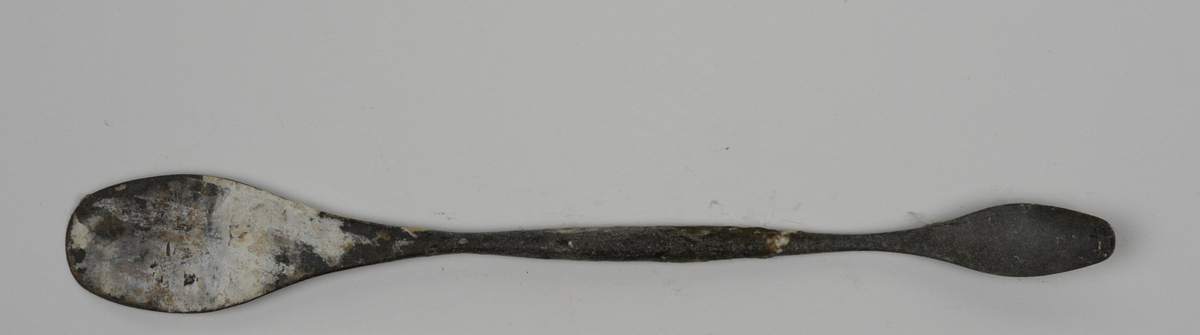 Modellerpinne i metall brukt av Nils Aas. Hver ende av pinnen er formet i en flat skje. Endene er noe bøyd i hver sin retning. Det er rester etter gips og annet materiale på verktøyet.