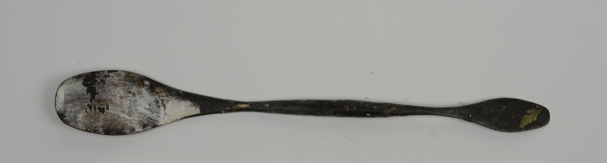 Modellerpinne i metall brukt av Nils Aas. Hver ende av pinnen er formet i en flat skje. Endene er noe bøyd i hver sin retning. Det er rester etter gips og annet materiale på verktøyet.