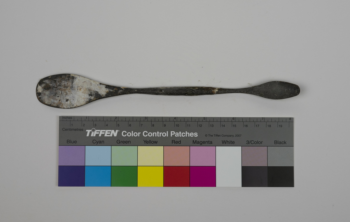 Modellerpinne i metall brukt av Nils Aas. Hver ende av pinnen er formet i en flat skje. Endene er noe bøyd i hver sin retning. Det er rester etter gips og annet materiale på verktøyet.