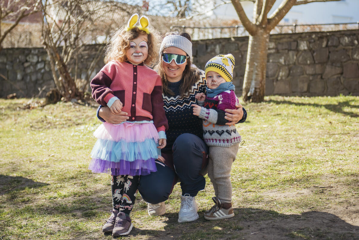 En dame med solbriller sitter på huk med to små barn på hver sin side. Det er vår og sol