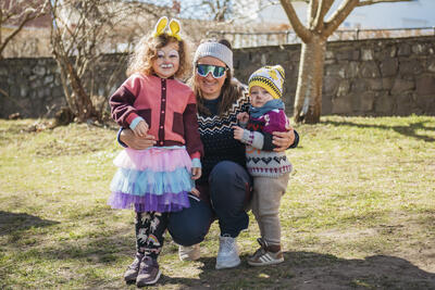 En dame med solbriller sitter på huk med to små barn på hver sin side. Det er vår og sol