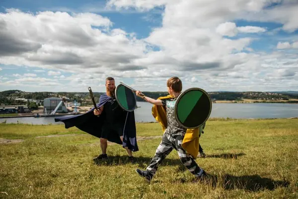 En voksen mann og en gutt ikledd middelalderdrakter kriger med sverd og skjold på toppen av et fjell