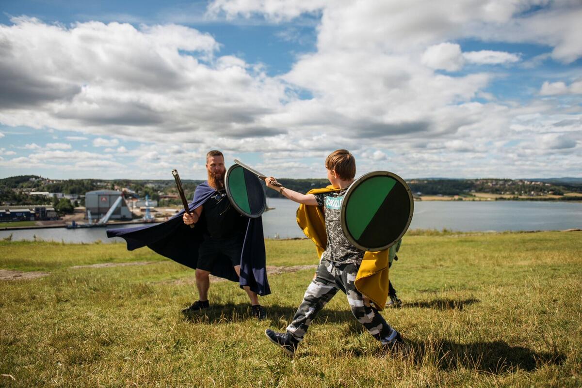 En voksen mann og en gutt ikledd middelalderdrakter kriger med sverd og skjold på toppen av et fjell