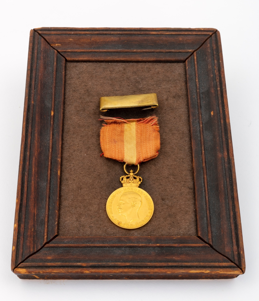 Kongens fortjenstmedalje i gull, med bånd i rødt med gul stripe i midten; alt festet til ramme av tre. På forsiden av medaljen er et portrett av Kong Haakon, omkranset av teksten "Haakon VII Norges Konge. Alt for Norge."