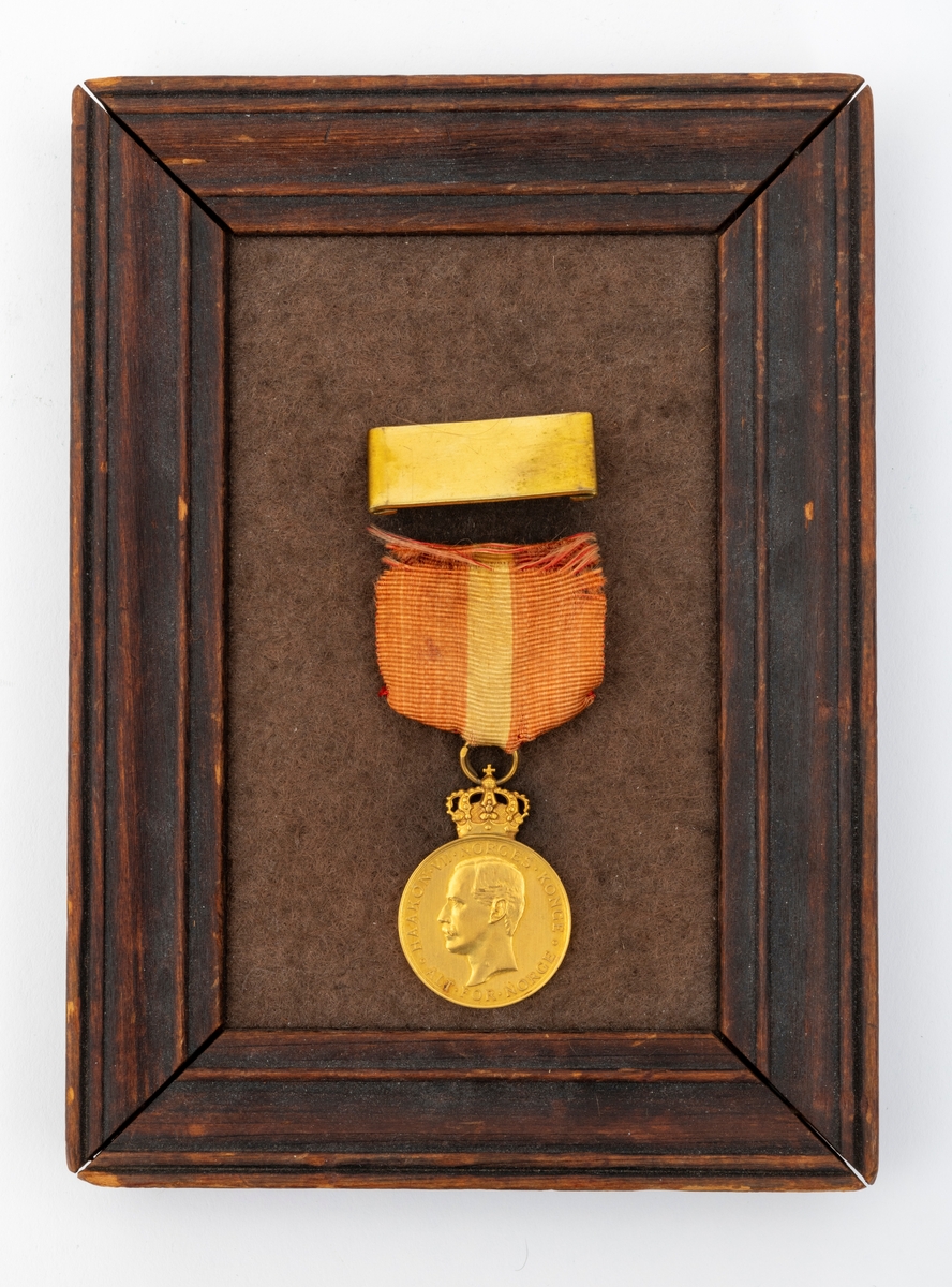 Kongens fortjenstmedalje i gull, med bånd i rødt med gul stripe i midten; alt festet til ramme av tre. På forsiden av medaljen er et portrett av Kong Haakon, omkranset av teksten "Haakon VII Norges Konge. Alt for Norge."