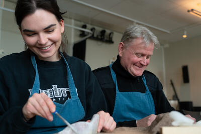 To personer, en dame og en mann sitter ved et bord og jobber med håndarbeid. De smiler
