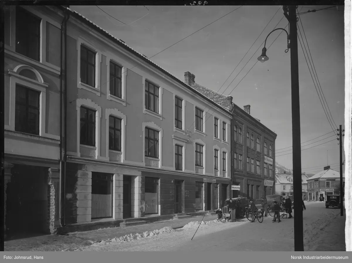 Bjørnstjerne Bjørnsons gate mot torget på vinterstid. Barn og voksne på sykkel og spark i gaten. Biler parkert langs fortau.