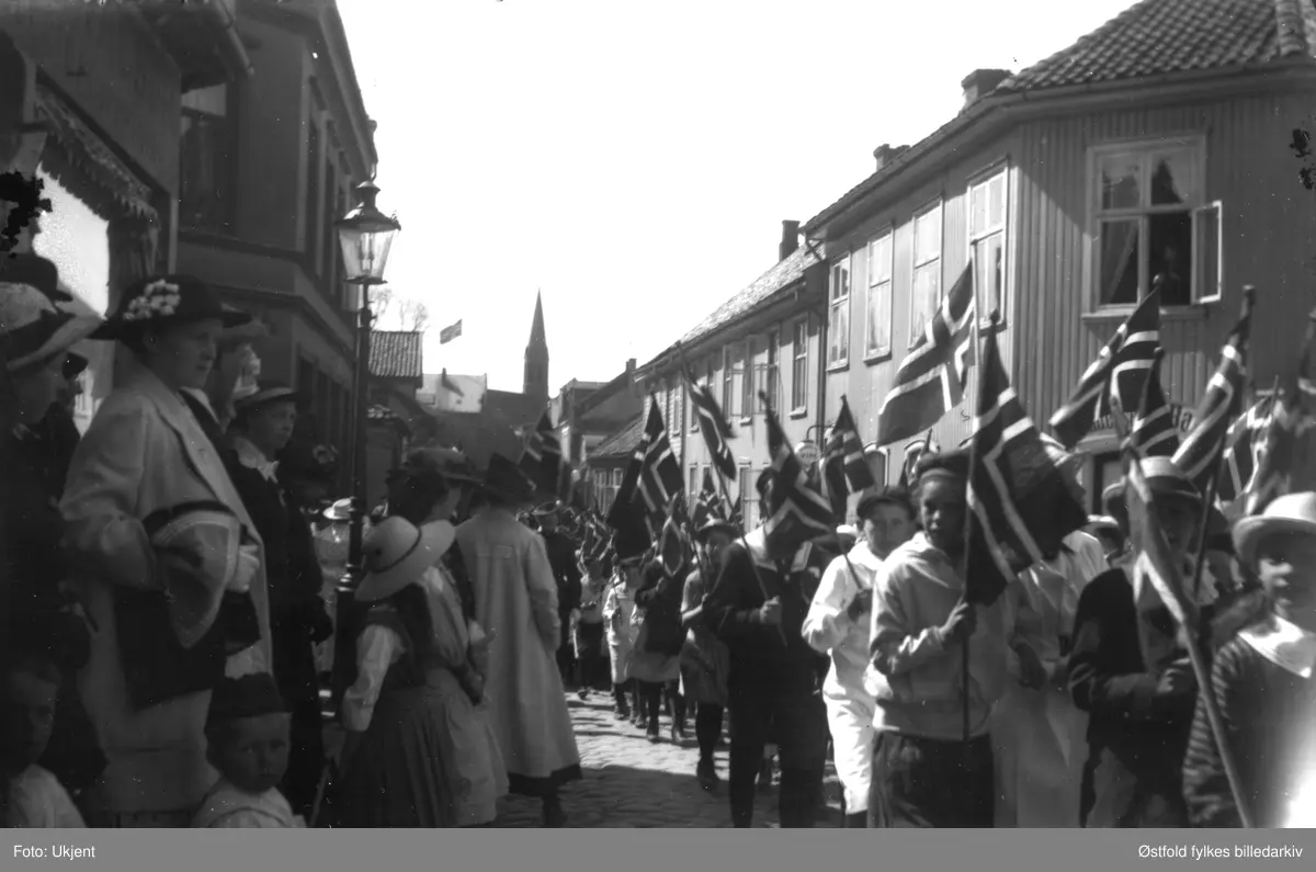 17. mai 1908 i Dronningensgate i Moss. Huset til høyre er Andersen & Baugs gård nr. 1. Huset (vinduet) til venstre er kjøpmann Walboms hus og forretning. Forretningen opphørte i 1926.