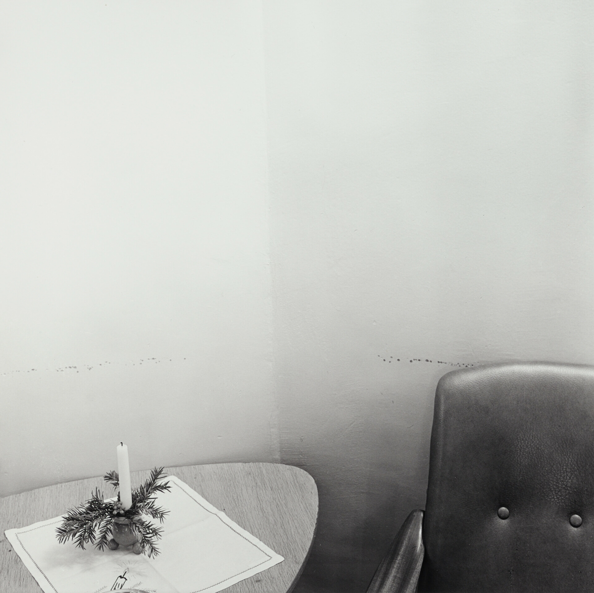 Et bord med juleduk og en pyntet lysestake med et stearinlys. En stol står ved siden av. De hvite veggene har merker.