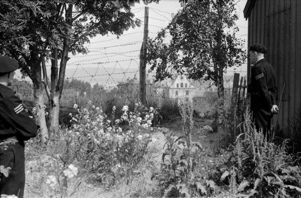 Flere menn, både i uniform og sivil står med ryggen til og ser mot et piggtrådgjerde.