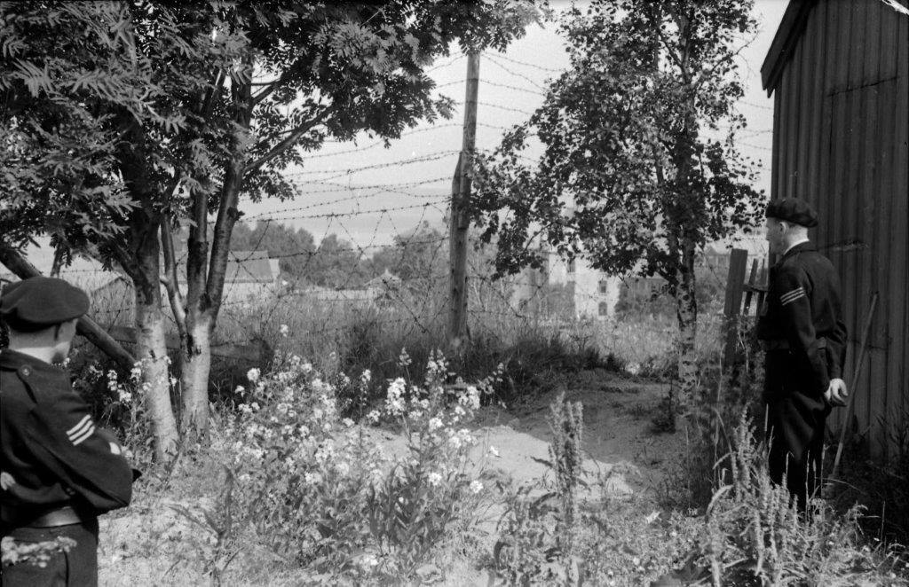 Flere menn, både i uniform og sivil står med ryggen til og ser mot et piggtrådgjerde.