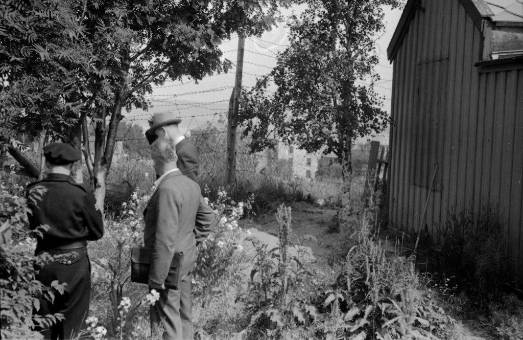 Flere menn, både i uniform og sivil står med ryggen til og ser mot et piggtrådgjerde.