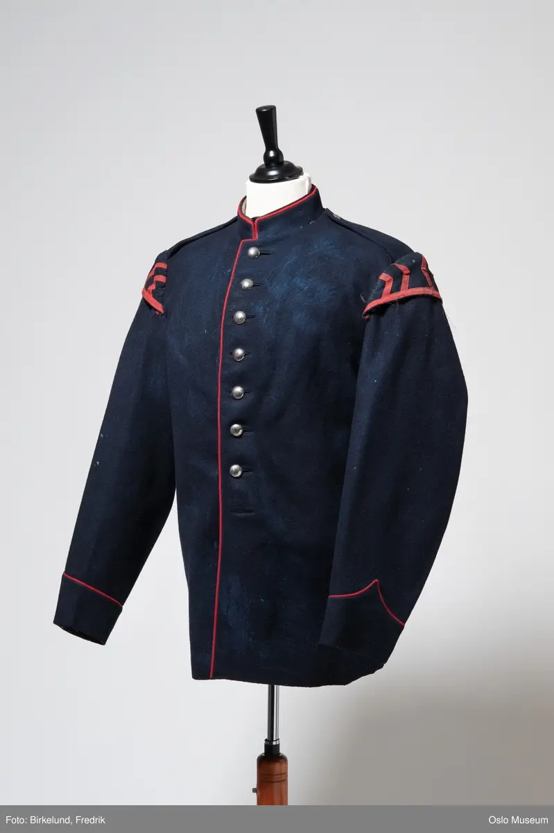 Uniformsjakke i sort, med røde kanter.
Omsydd for bruk som kostyme ved Det Norske Teater: det er satt inn puffer på armene.